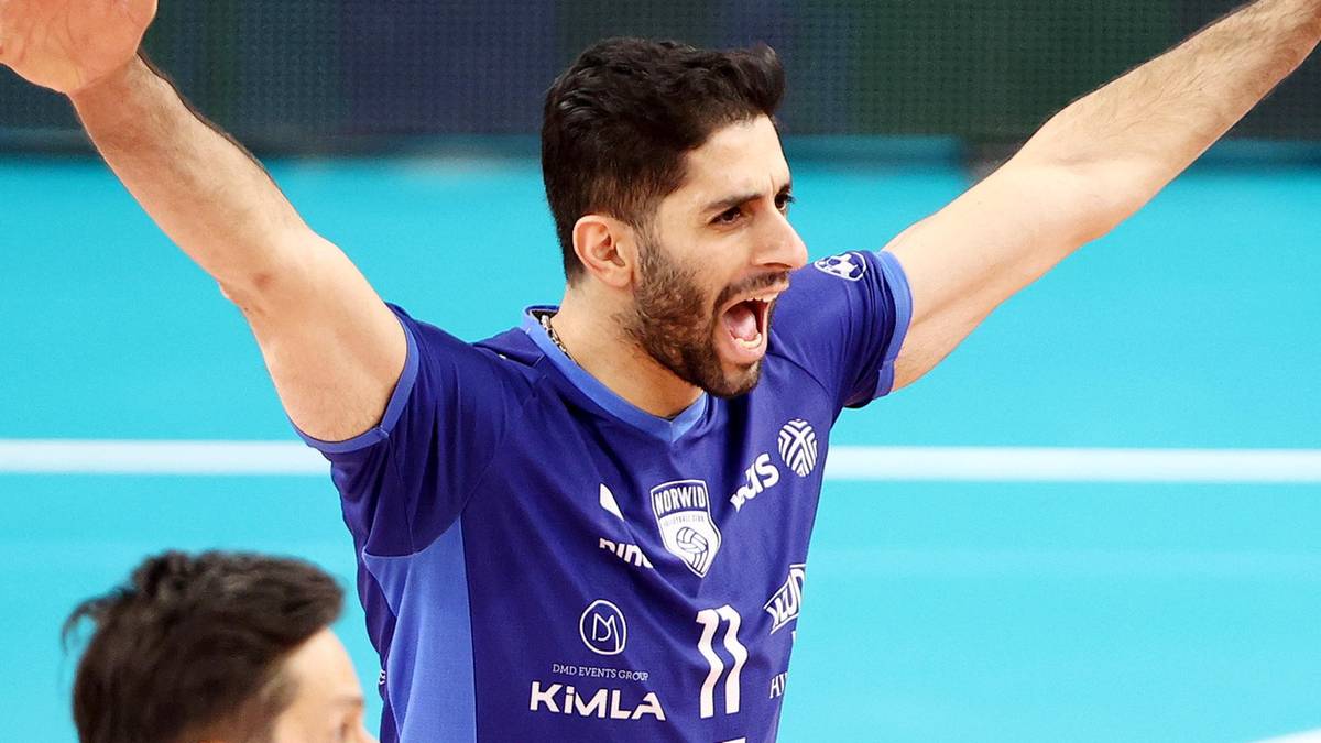 Siatkarz w niebieskiej koszulce, patrzący w bok z uniesionymi ramionami i otwartymi ustami.