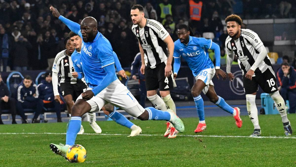 Piłkarz w niebieskiej koszulce uderza piłkę nogą, jego ciało jest wygięte do przodu, podczas meczu piłki nożnej. W tle widać innych graczy w białych i czarnych koszulkach oraz widzów na trybunach.