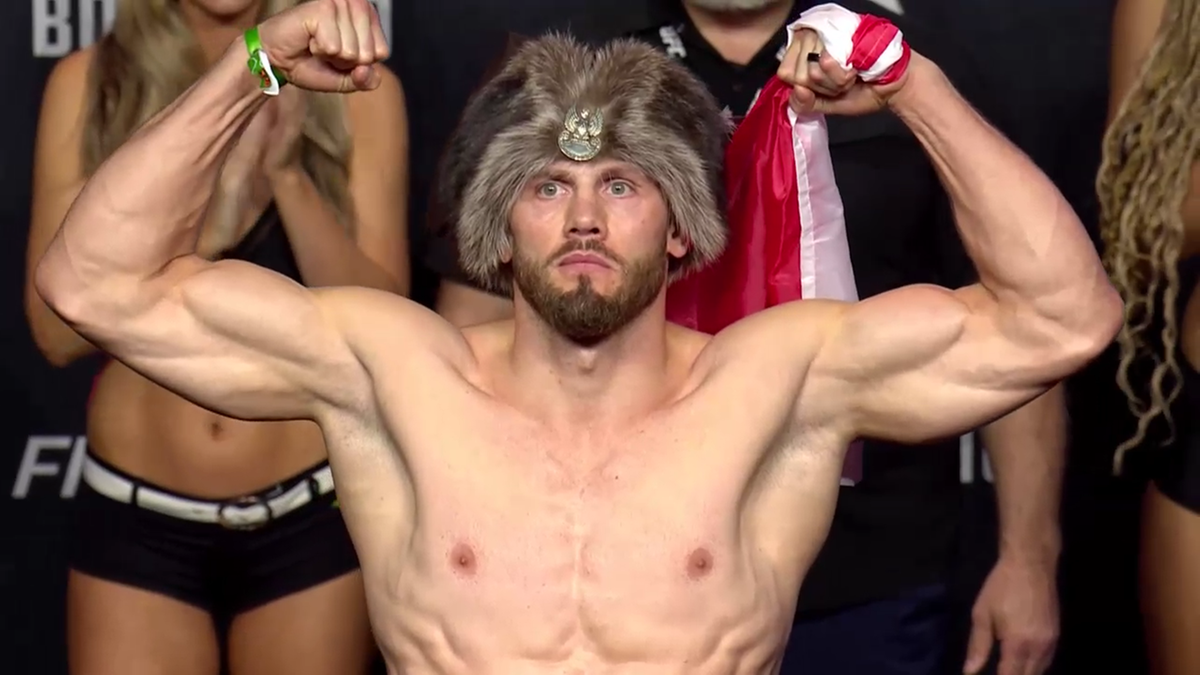 Mężczyzna z wąsami i brodą, ubrany w futrzaną czapkę z orłem, napina bicepsy i pokazuje mięśnie klatki piersiowej. W tle widać fragmenty innych osób i chorągiew Polski.