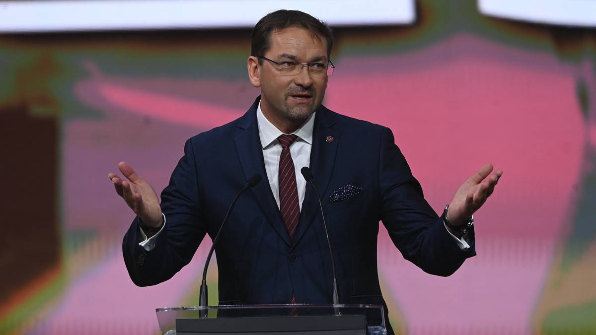 Mężczyzna w ciemnym garniturze i czerwonym krawacie, stojący za mównicą, gestykulujący rękami.