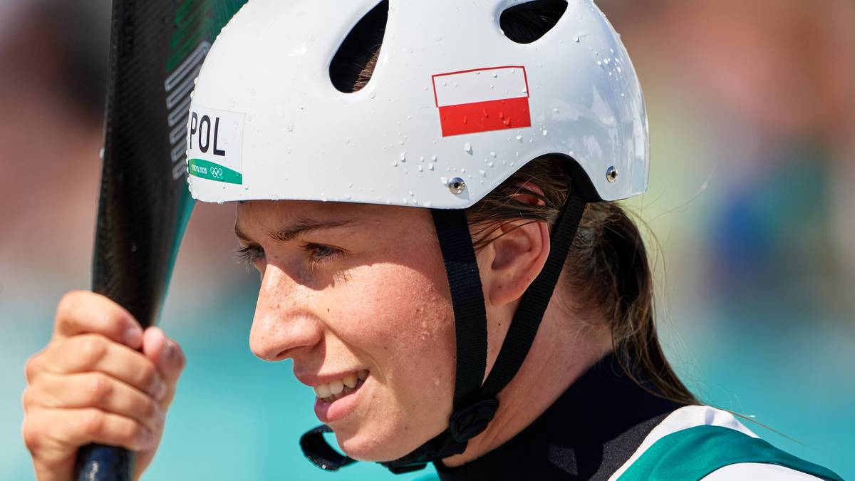 Kobieta w białym kasku z polską flagą, przygotowuje się do startu w zawodach kajakowych.