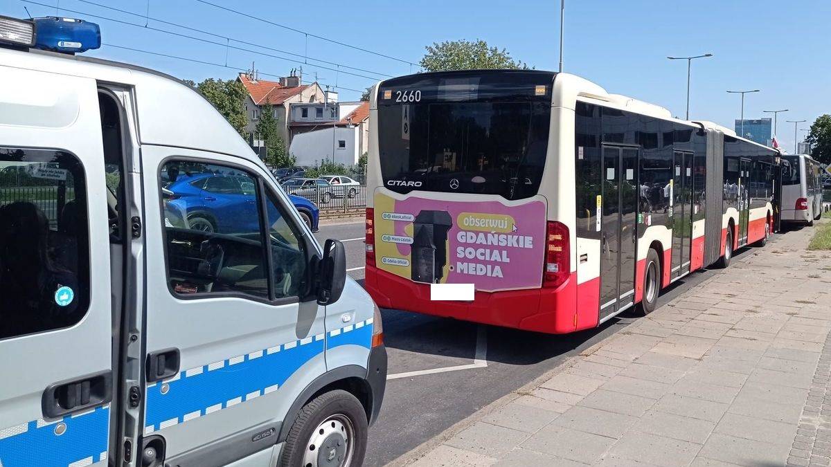 Dwa autobusy stojące na ulicy po kolizji. Widać fragment białego furgonu z niebieskimi pasami po lewej stronie. Dalej, na drodze, znajdują się dwa przegubowe autobusy, jeden z nich ma czerwony dół i białą górę z napisem "GDAŃSKIE SOCIAL MEDIA". W tle widać budynki i drzewa.
