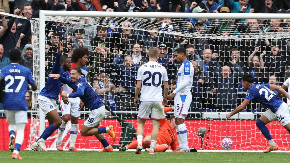 Piłkarze w niebieskich strojach świętują zdobycie bramki w meczu piłki nożnej, podczas gdy zawodnicy w białych strojach wydają się zawiedzeni. Na trybunach widać kibiców.