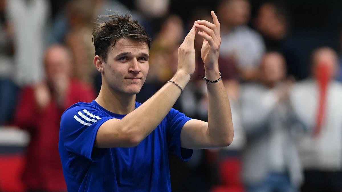 Młody mężczyzna w niebieskiej koszulce z białymi pasami na ramionach, z dłońmi złożonymi do oklasków.