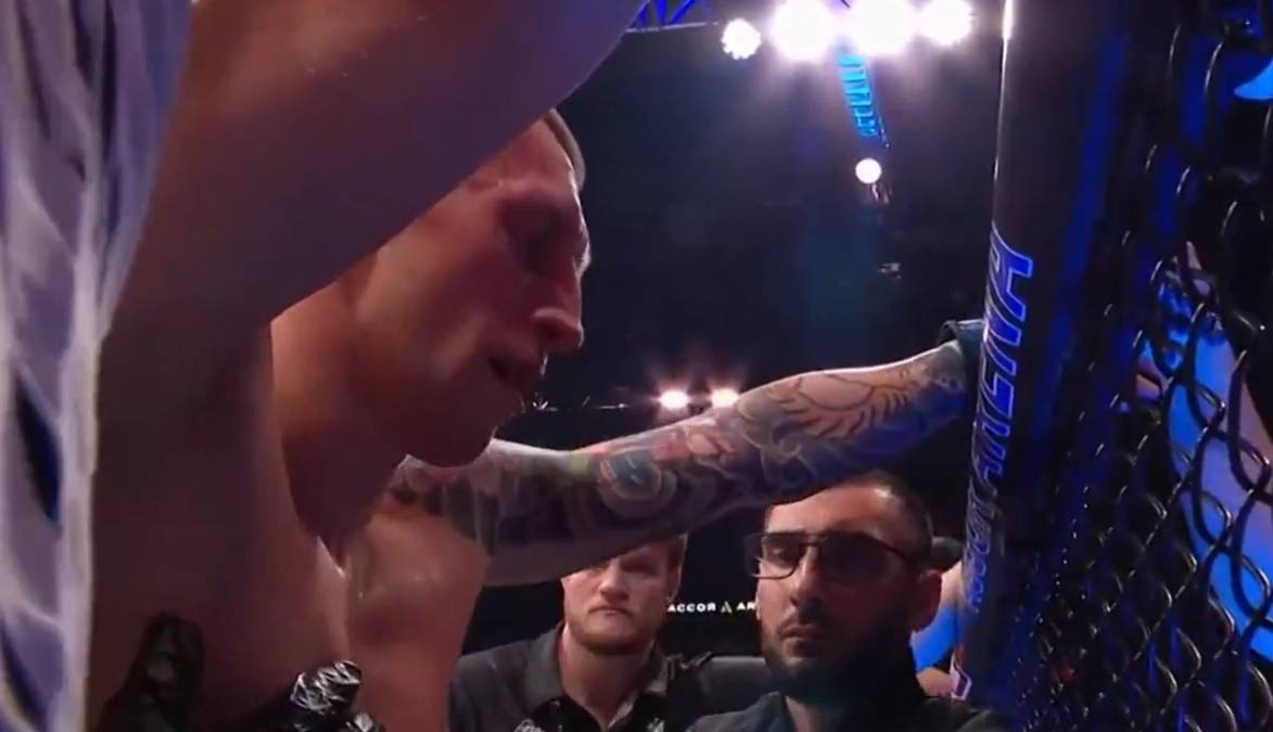 Wojownik UFC z tatuażami na ramieniu nachyla się nad klatką, w tle widoczni są inni mężczyźni.