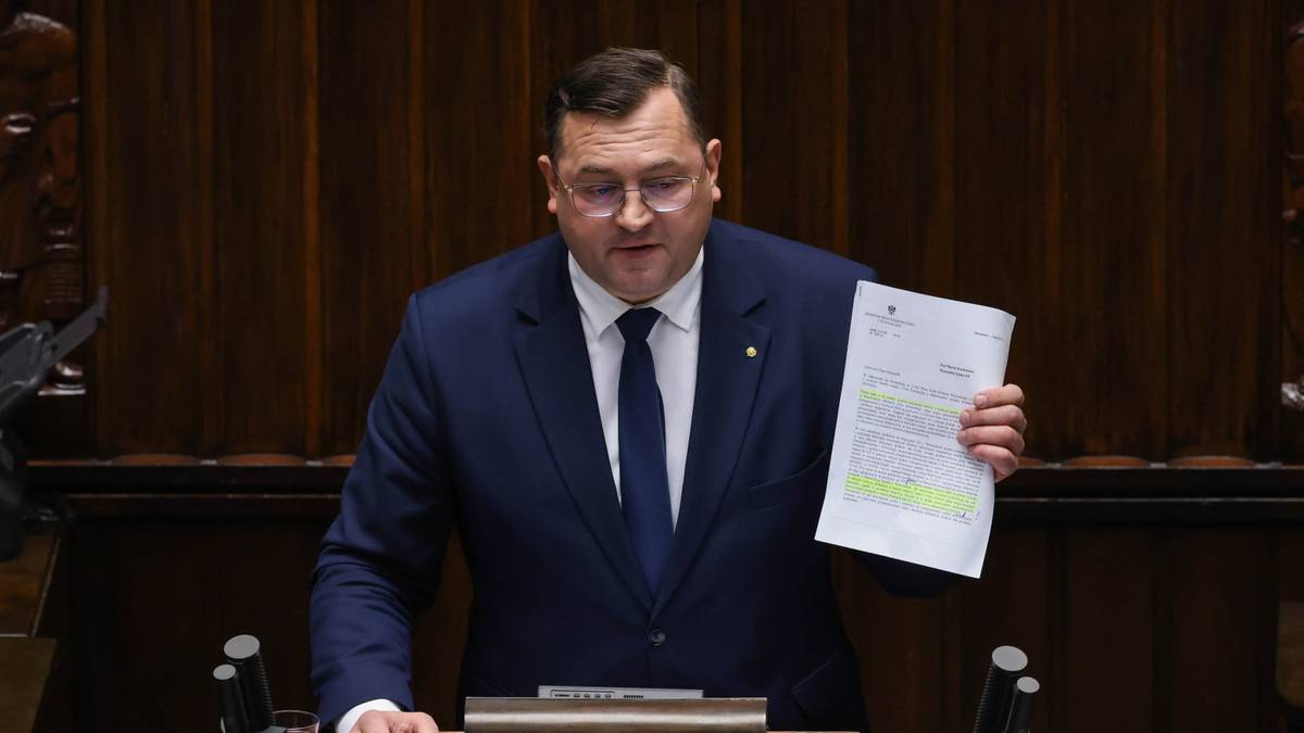 Polityk w garniturze przemawia za m&oacute;wnicą, trzymając dokument w ręku.