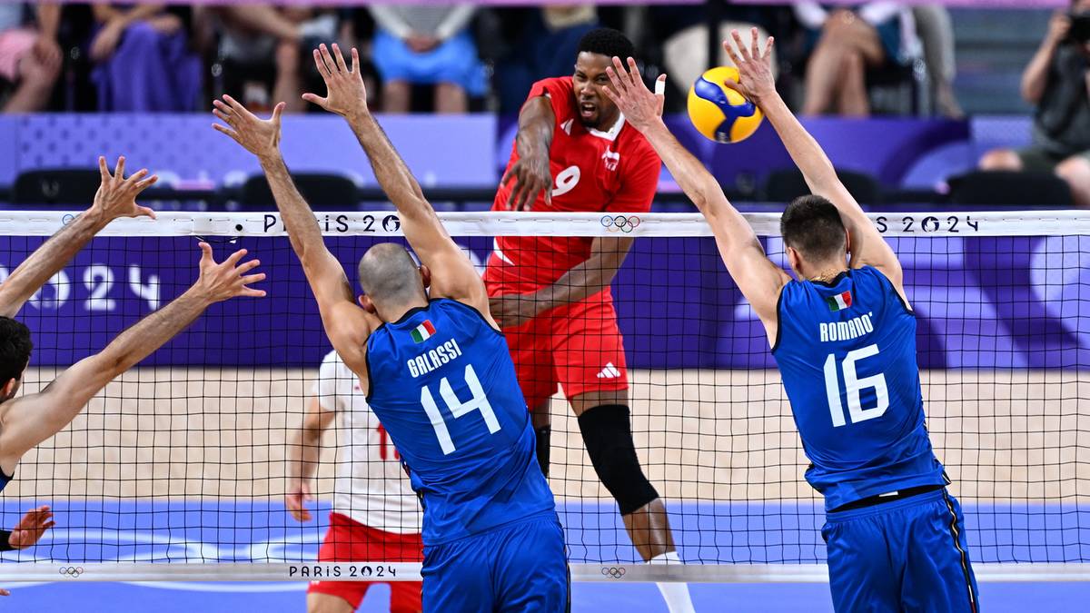 Siatkarze rywalizujący podczas meczu. Jeden z zawodników z drużyny w niebieskich strojach próbuje zablokować atak przeciwnika w czerwonej koszulce.