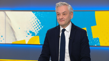 Mężczyzna w białej koszuli i krawacie siedzi w studiu telewizyjnym, uśmiechając się.