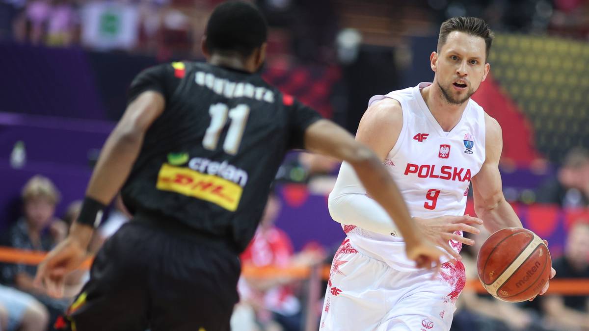 Koszykarz w białej koszulce z napisem Polska i numerem 9 na plecach, biegnie z piłką w ręku, broniony przez przeciwnika w czarnej koszulce z numerem 11.