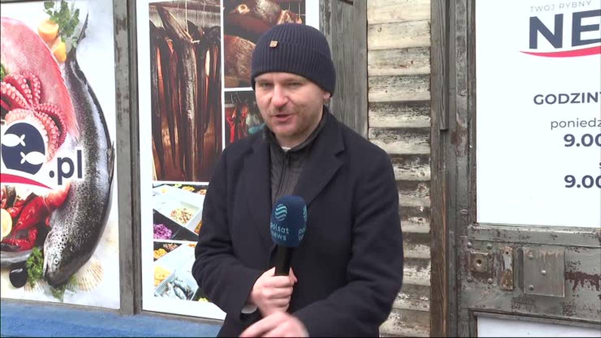 Mężczyzna z mikrofonem przed ustami, ubrany w czapkę i płaszcz, stoi na tle plakatu z rybami i szyldu sklepu.