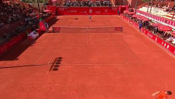 Widok z g&oacute;ry na kort tenisowy z czerwonej mączki, na kt&oacute;rym grają dwaj tenisiści. Na trybunach siedzą widzowie, a w tle widać reklamę ATP.