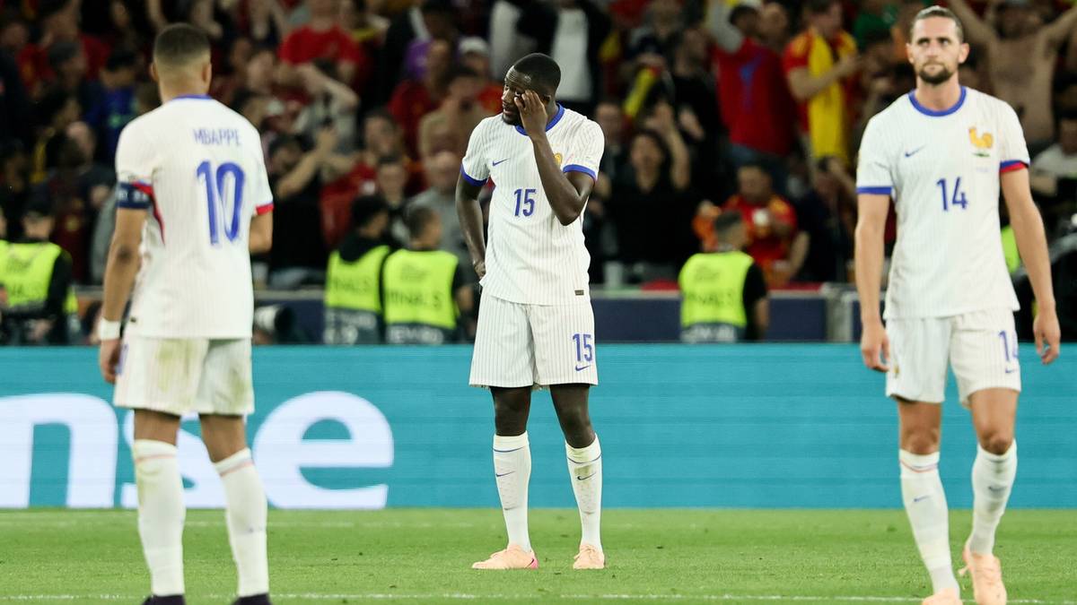 Trzech piłkarzy w białych koszulkach i białych spodenkach stoi na boisku piłkarskim.