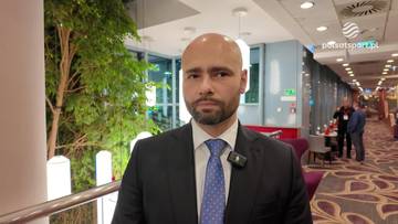 Mężczyzna w garniturze i krawacie, z krótką brodą i łysą głową, patrzy prosto w obiektyw. Na jego klapie marynarki znajduje się mikrofon.