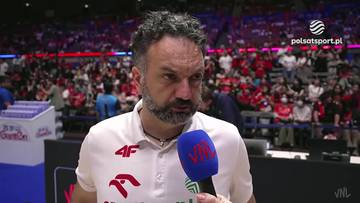 Stefano Lavarini, trener siatk&oacute;wki, w białej koszulce z logo 4F i polskimi orzełkami, trzymający mikrofon podczas wywiadu. W tle widoczna jest publiczność na trybunach.