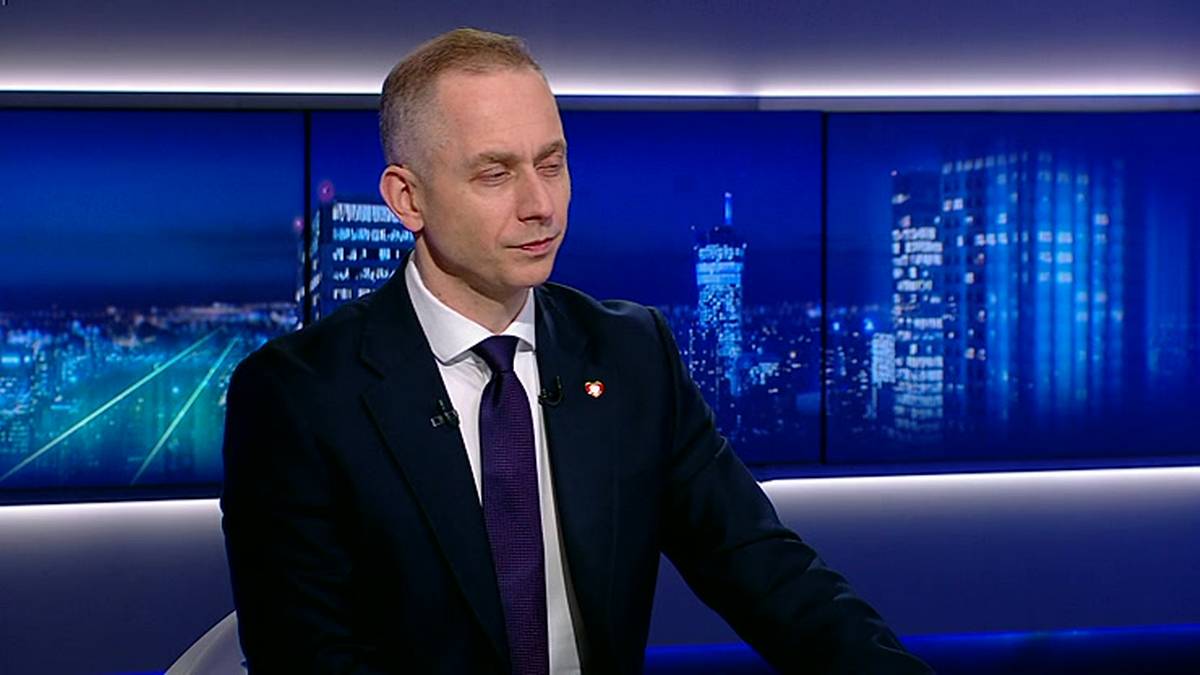 Mężczyzna w ciemnym garniturze i fioletowym krawacie siedzi na krześle. Za nim znajduje się niebieski ekran z podświetlonymi sylwetkami budynk&oacute;w.