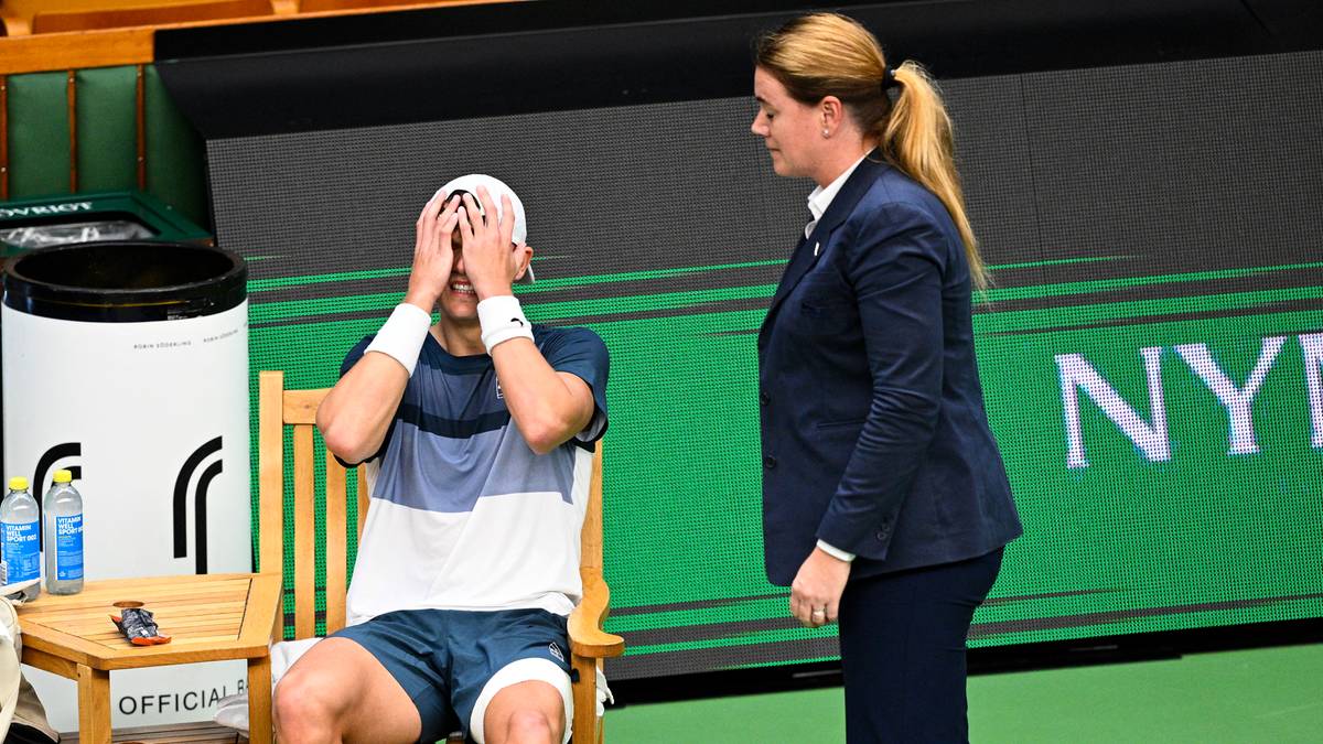 Tenisista siedzi na krześle z rękami na twarzy, wyrażając smutek lub ból. Obok stoi kobieta w stroju sportowym.