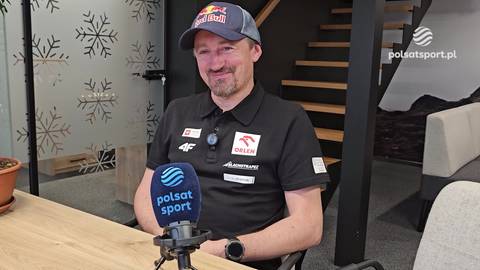 Adam Małysz w czapce z daszkiem i koszulce z logo sponsora, siedzi przed mikrofonem Polsat Sport.