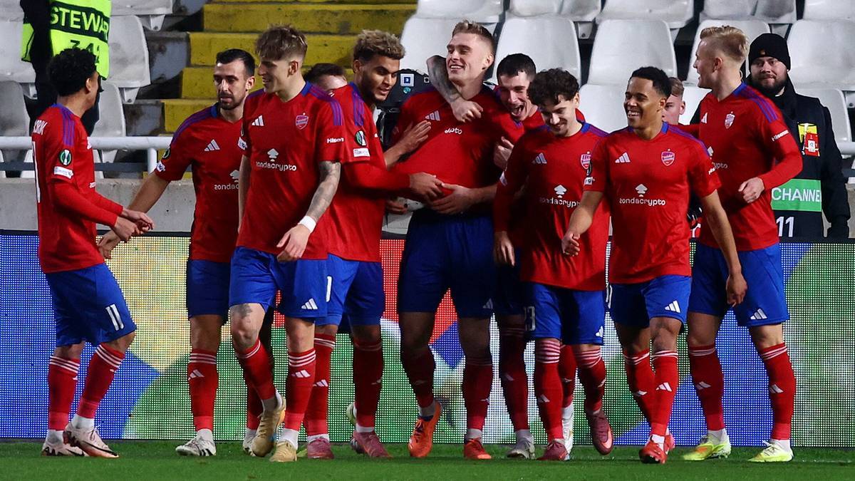 Drużyna piłkarska w czerwonych strojach świętuje gola, wielu zawodnik&oacute;w przybija sobie piątki i obejmuje się.