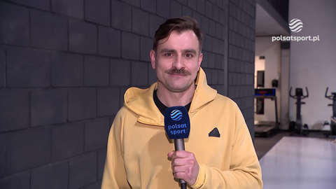 Jan Szymański w ż&oacute;łtej bluzie z kapturem, z wąsami, trzyma mikrofon Polsat Sport. W tle widać bieżnię i inne urządzenia sportowe.