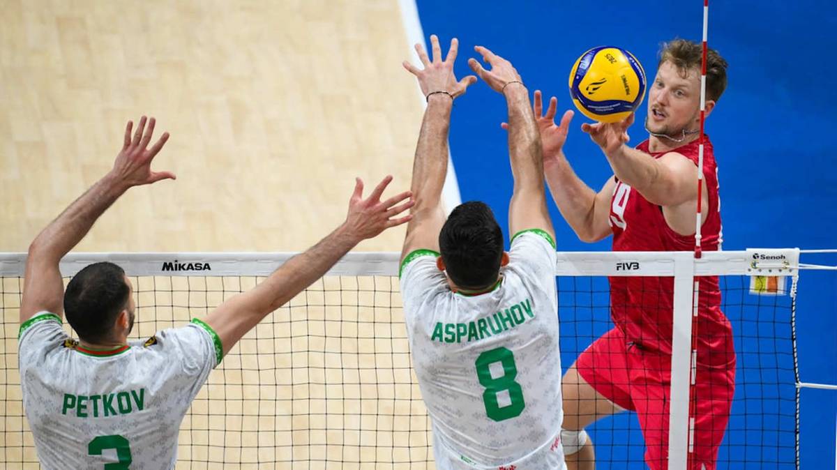 Siatkarz w czerwonej koszulce atakuje piłkę, podczas gdy dwaj gracze w białych koszulkach blokują go.