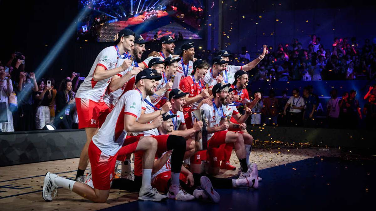 Drużyna siatkarzy w strojach biało-czerwonych świętuje zwycięstwo, część z nich siedzi na ziemi, inni stoją wokół.