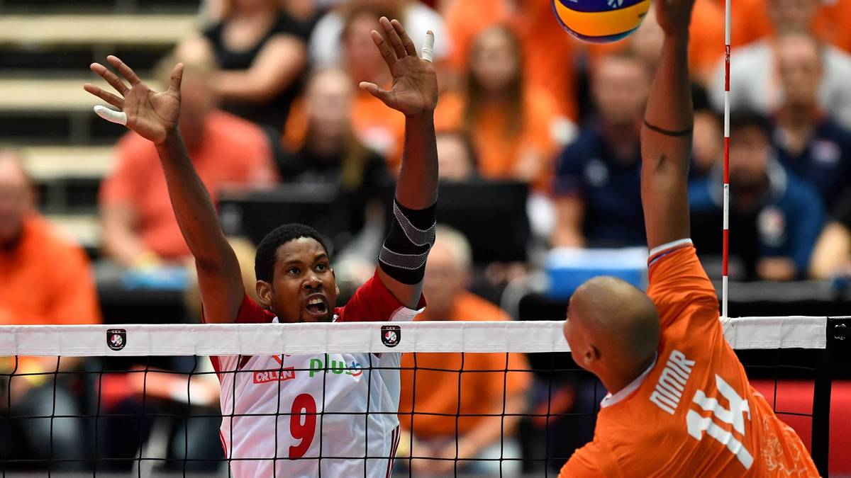 Dwóch siatkarzy blokuje piłkę nad siatką. Jeden z nich, w białej koszulce z numerem 9, wyskakuje w górę z otwartymi dłońmi. Drugi, w pomarańczowej koszulce z numerem 14, również próbuje zatrzymać piłkę.
