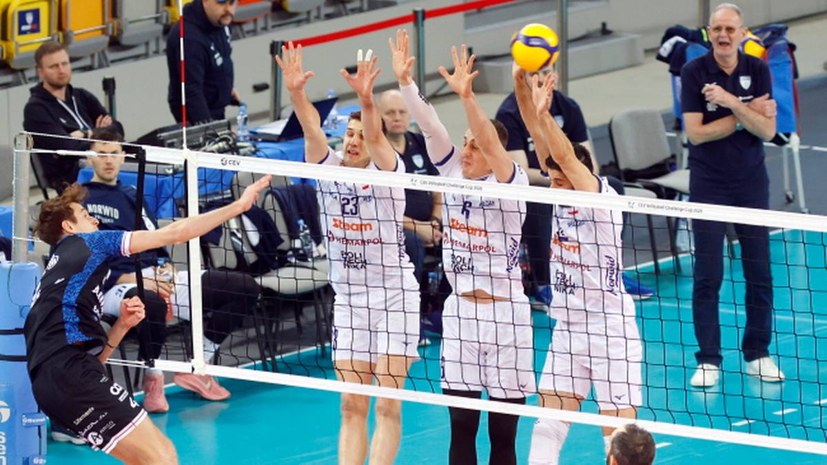 Siatkarze w białych strojach blokują atak przeciwnika podczas meczu siatkówki.