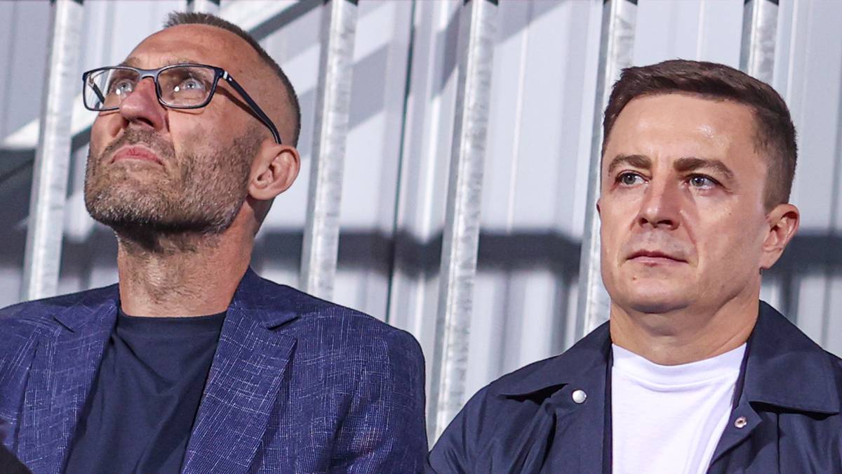 Trzech mężczyzn siedzi obok siebie, dw&oacute;ch w garniturach i jeden młodszy w szaliku.