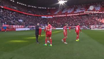 Piłkarze w czerwonych strojach stoją na zielonej murawie stadionu, w tle widać trybuny wypełnione kibicami w czerwonych barwach.