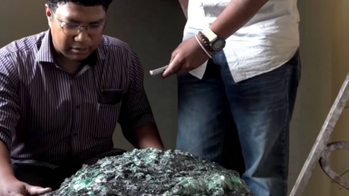 Mężczyzna w okularach pochyla się nad ciemnym, pokruszonym kamieniem z zielonymi kryształami, obok stoi druga osoba.