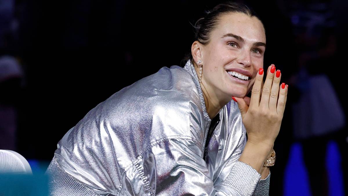 Aryna Sabalenka, tenisistka, w błyszczącej, srebrnej kurtce, uśmiecha się szeroko, patrząc w bok.