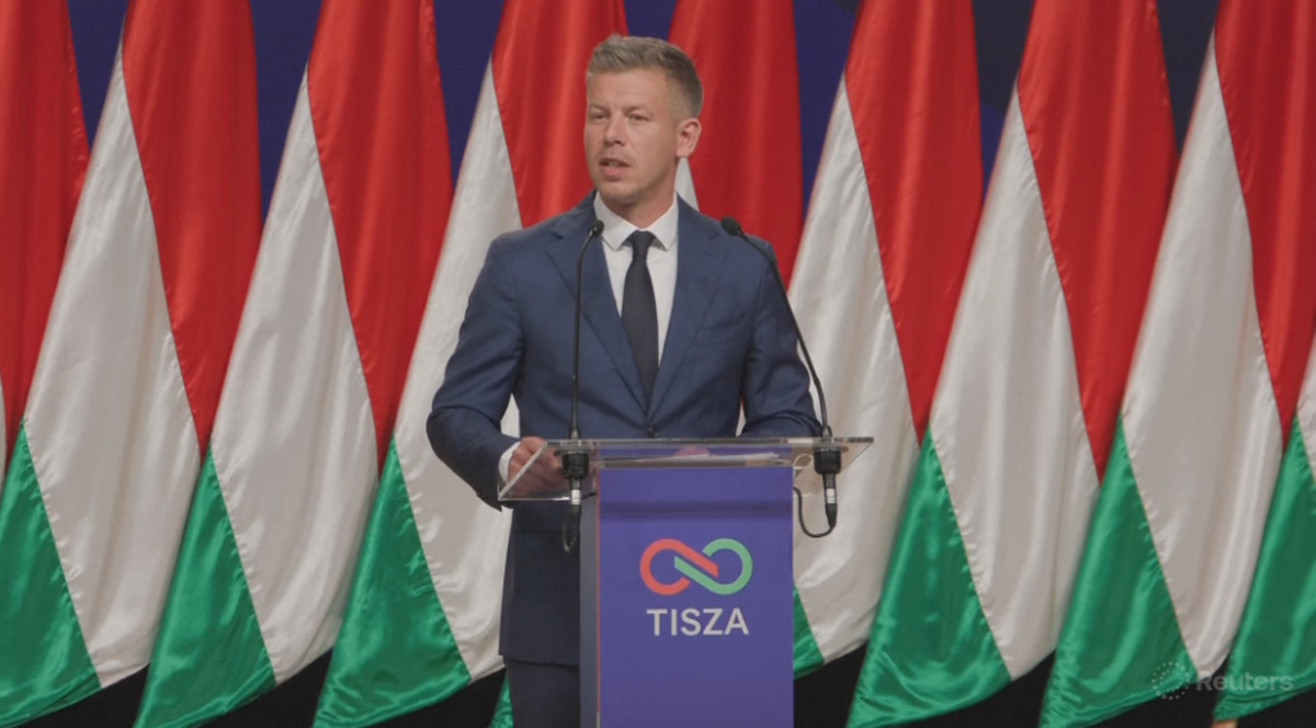 Peter Magyar stoi przy m&oacute;wnicy na tle flag Węgier, przemawia publicznie.