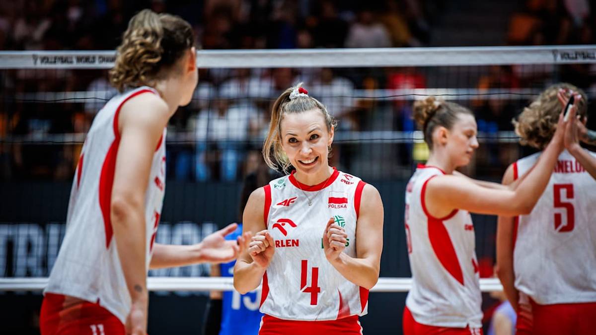 Siatkarka z numerem 4 na koszulce, z krótkimi blond włosami, uśmiecha się w stronę kamery. Jest w białej koszulce z czerwonymi elementami i numerem 4. Wokół niej widać inne zawodniczki w podobnych strojach, a w tle trybuny z kibicami.