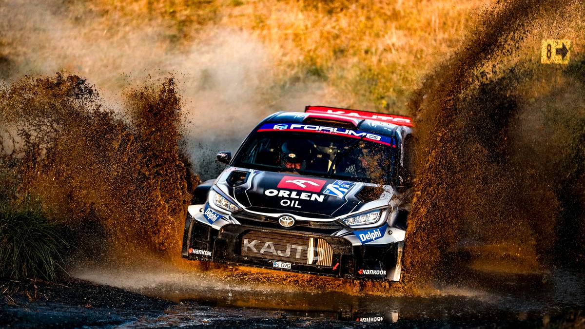 Samochód rajdowy z widocznym napisem "KAJTO" na przedniej szybie, jadący przez głęboką kałużę błota, rozpryskując wodę wokół.