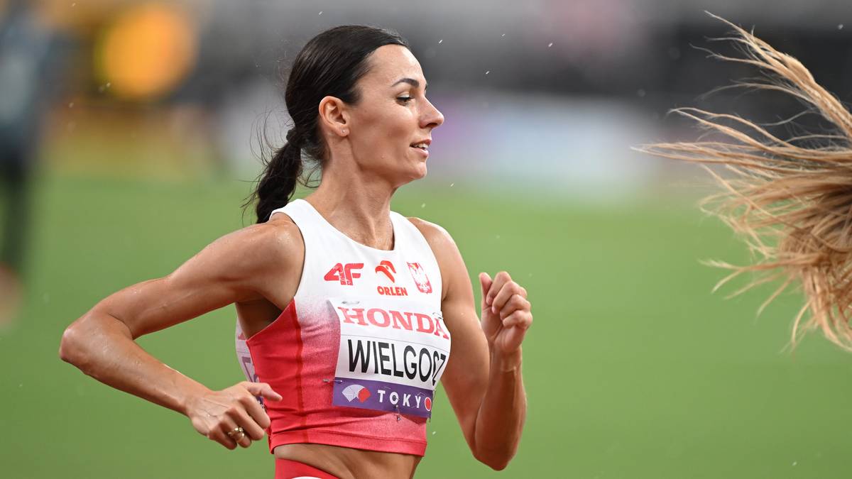 Lekkoatletka podczas biegu, w białej koszulce z czerwonymi elementami i napisem "WIELGÓR" oraz "TOKYO". Kobieta ma spięte włosy i koncentruje się na biegu. Jest lekko pochylona do przodu.
