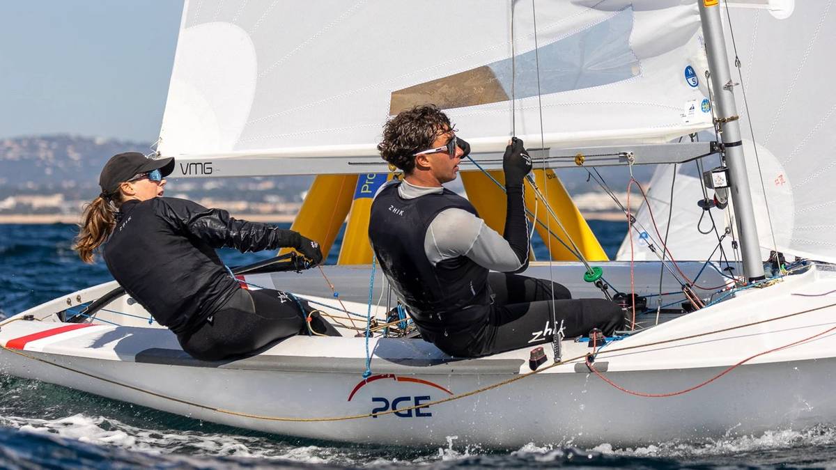 Dwoje ludzi na żagl&oacute;wce, jeden steruje, drugi obsługuje żagiel. Osoby ubrane w stroje sportowe, na wodzie.