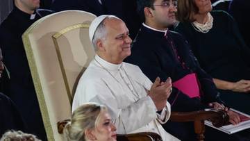 Papież Leon XIV siedzi w fotelu, z rękami złożonymi w geście aplauzu, otoczony przez duchownych i świeckich.