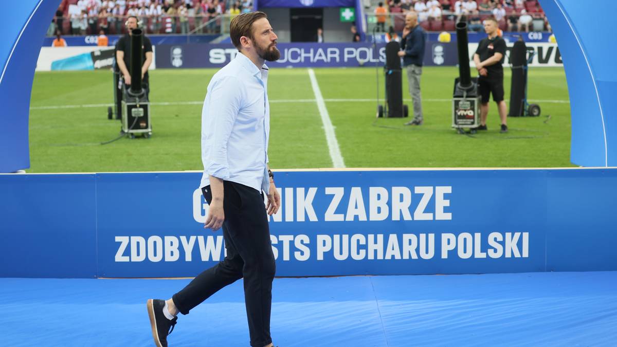 Mężczyzna w jasnej koszuli i czarnych spodniach idzie po niebieskim dywanie. W tle widać murawę stadionu piłkarskiego z widzami na trybunach.