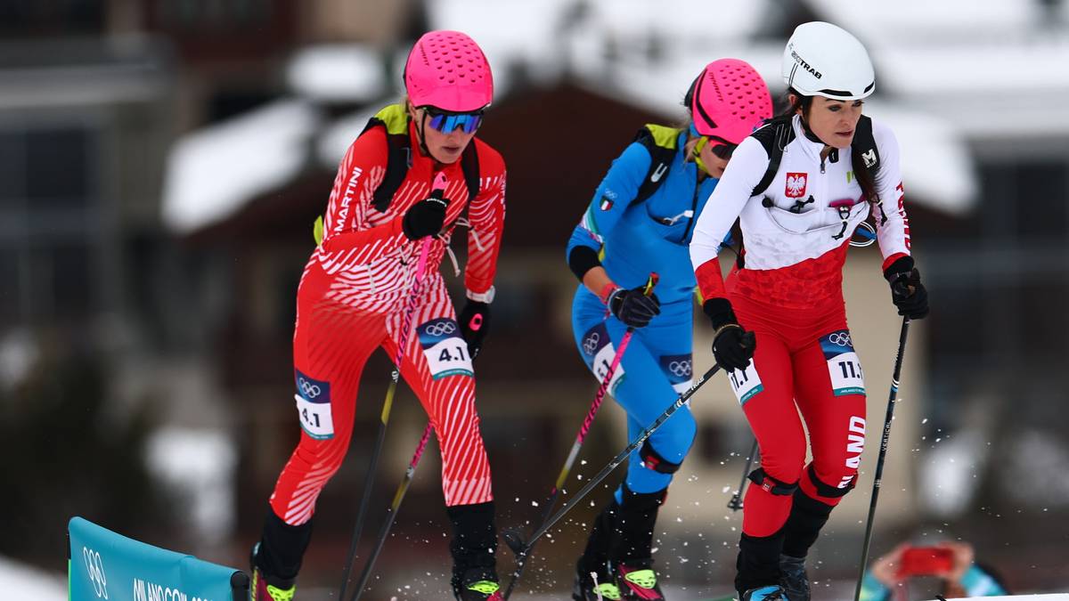 Trzy zawodniczki w strojach narciarskich, z kaskami i kijkami, rywalizują w zawodach skialpinistycznych na ośnieżonym stoku.