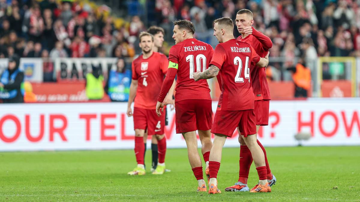 Piłkarze reprezentacji Polski w czerwonych strojach świętują zwycięstwo na boisku, jeden z nich wskazuje w kierunku publiczności.