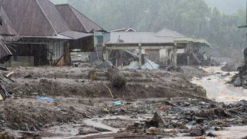 Budynki zniszczone przez powódź i osuwisko, zalanymi błotem i gruzem, w tle wzgórza pokryte zielenią.