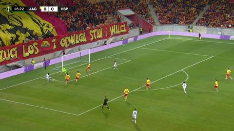 Piłkarz w zielonej koszulce wykonuje paradę bramkarską, broniąc strzał w meczu piłki nożnej.