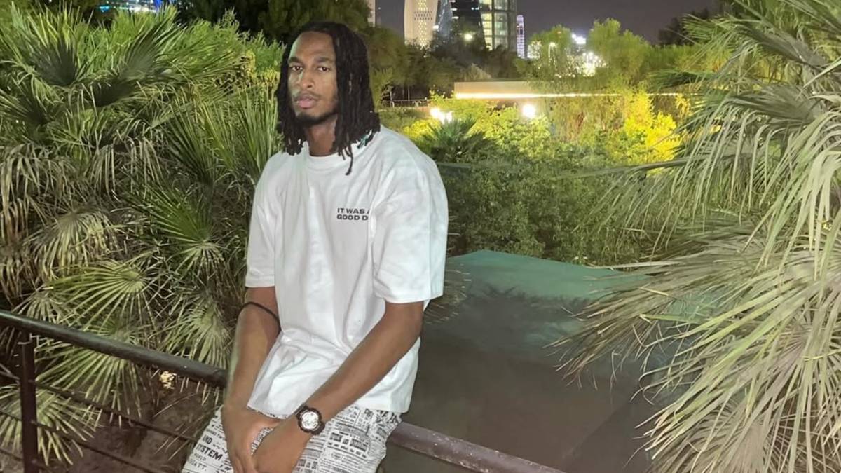 Młody mężczyzna o ciemnej karnacji i długich warkoczach w białej koszulce z krótkim rękawem i zegarkiem na nadgarstku, siedzi na balustradzie na tle nocnej roślinności.