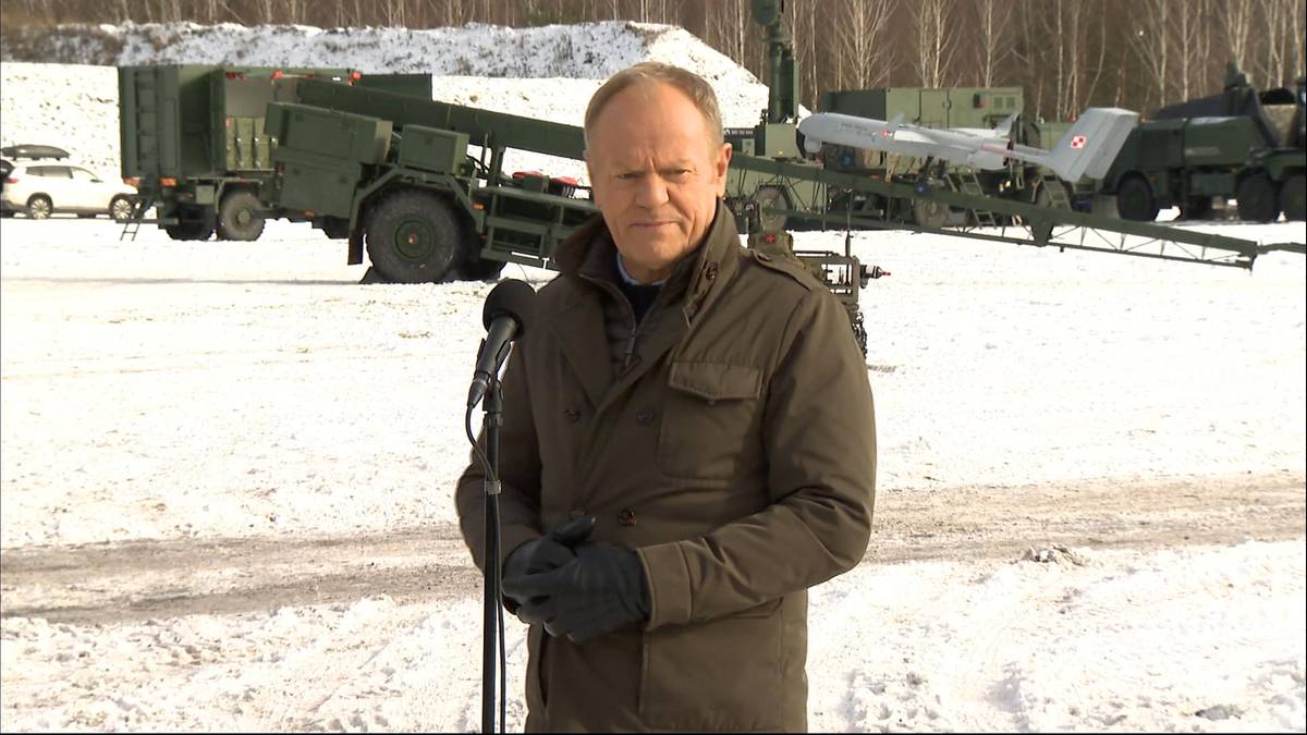 Premier Donald Tusk przemawia przed mikrofonem, w tle widoczne wozy bojowe i zaśnieżony teren.