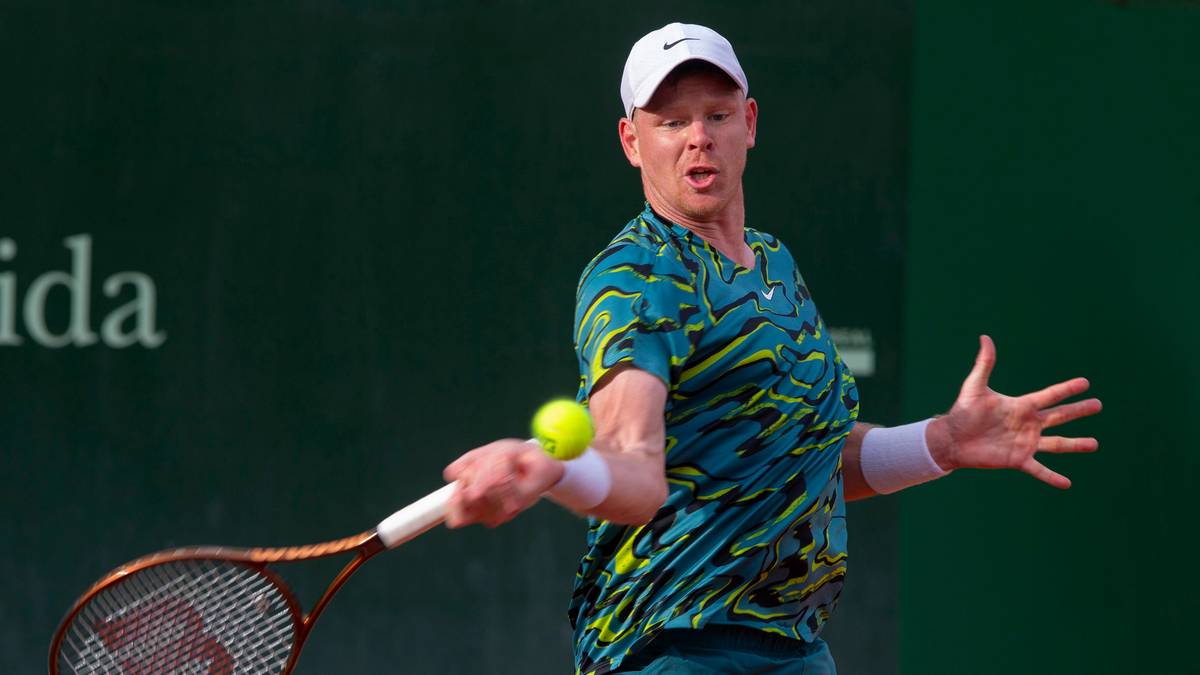 Tenisista z rudymi włosami, w zielono-niebieskiej koszulce i białej czapce, wykonuje forhendowy uderzenie podczas meczu tenisowego.