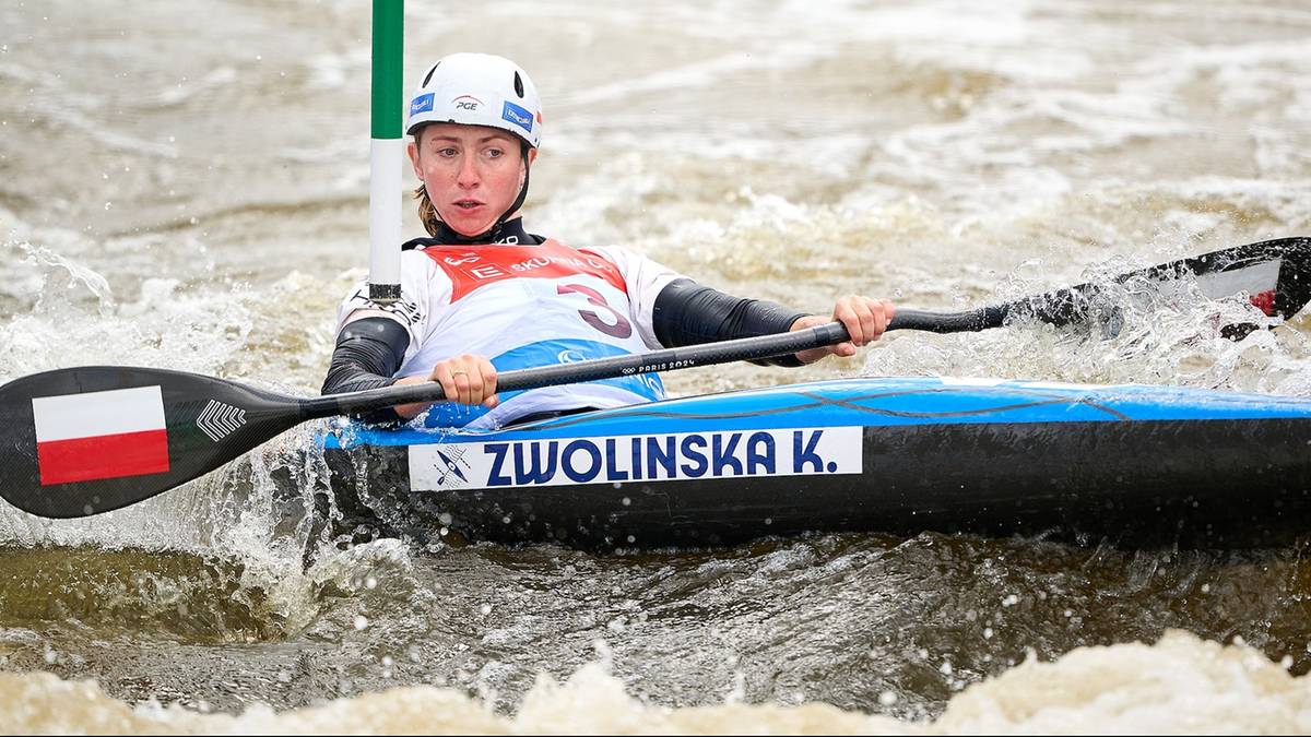 Kobieta w kasku i kamizelce ratunkowej wiosłuje w kajaku po wzburzonej wodzie. Na kajaku widnieje nazwisko "ZWOLINSKA K." oraz polska flaga.