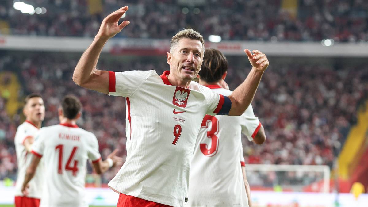 Piłkarz Robert Lewandowski w białej koszulce z orzełkiem i numerem 9 świętuje bramkę z podniesionymi rękami, na tle innych graczy w biało-czerwonych strojach i trybun wypełnionych kibicami.