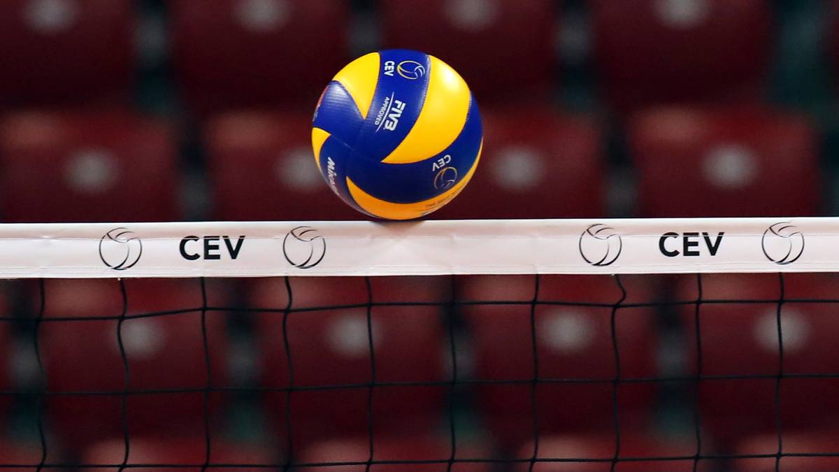 Piłka do siatkówki w kolorach żółtym i niebieskim leży na siatce do siatkówki. W tle widać czerwone krzesełka.