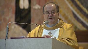 Uśmiechnięty mężczyzna w szatach liturgicznych stoi za mównicą z mikrofonem.