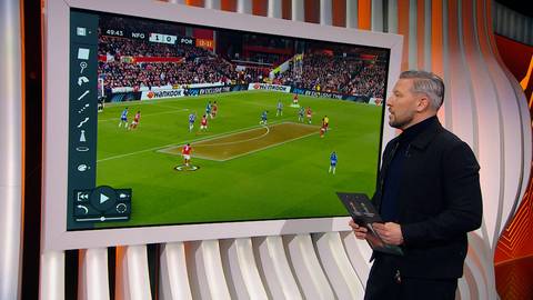 Mężczyzna w czarnym ubraniu stoi obok dużego ekranu, na kt&oacute;rym wyświetlany jest fragment meczu piłki nożnej między Nottingham Forest a FC Porto. Na ekranie widoczni są piłkarze na boisku.